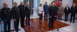 Przed mikrofonem przemawia oficer Państwowej Straży Pożarnej w mundurze galowym. W tle ustawieni w szeregu duchowni PSP, osoby w garniturach oraz przedstawiciele innych służb mundurowych.