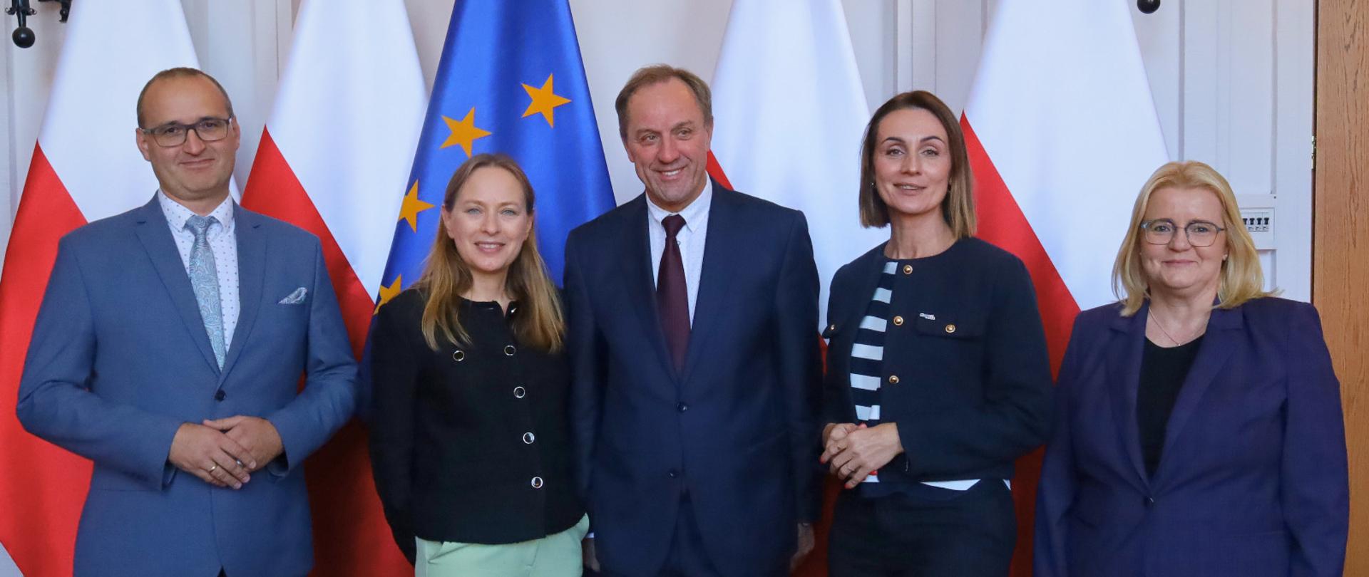 kilka osób - 3 kobiety i dwóch mężczyzn stoi na tle flag polskich i unijnych