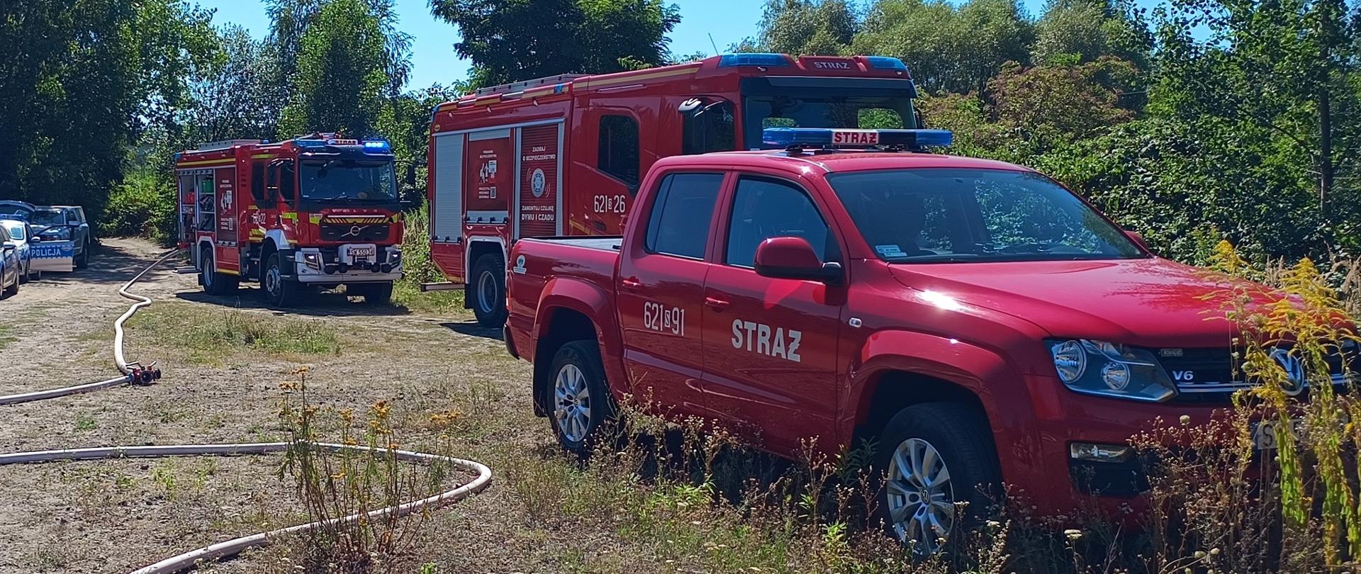 Na zdjęciu widoczne są strażackie pojazdy bojowe ustawione wzdłuż gruntowej drogi podczas ćwiczeń. Na pierwszym planie po prawej stronie zaparkowany jest czerwony pickup marki Volkswagen Amarok z napisem "STRAŻ", natomiast za nim stoi duży, ciężki wóz gaśniczy marki Volvo oraz kolejny pojazd pożarniczy na dalszym planie. Wzdłuż drogi przeciągnięte są rozwinięte węże strażackie.
Cała akcja odbywa się na otwartym, słonecznym terenie otoczonym gęstą roślinnością i drzewami. Po lewej stronie, w oddali za wozami strażackimi, widoczny jest również zaparkowany radiowóz policyjny.