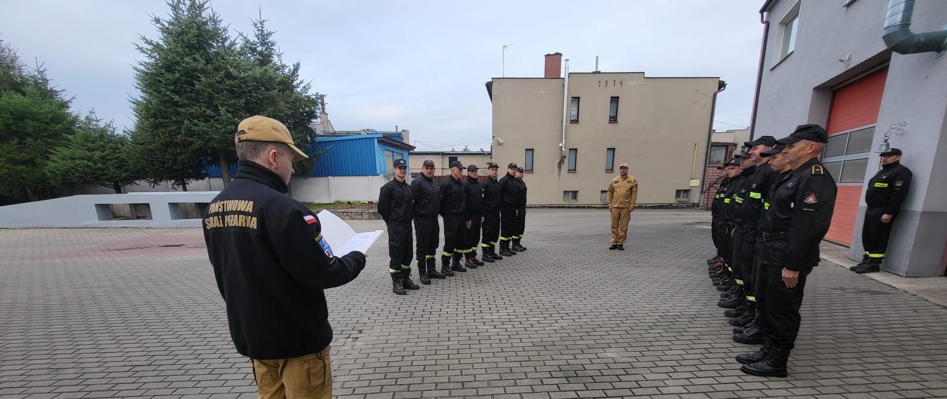 Komendant Powiatowy Państwowej Straży Pożarnej starszy brygadier Maciej Suchomski ubrany w mundur służbowy stoi naprzeciw Zastępcy Jednostki Ratowniczo Gaśniczej starszego kapitana Krzysztofa Brzozowskiego trzymając w rękach otwarte pismo rozkazu. Po jego lewej i prawej stronie w szeregu ubrani w mundury koszarowe stoją na baczność naprzeciw siebie strażacy dwóch zmian służbowych.