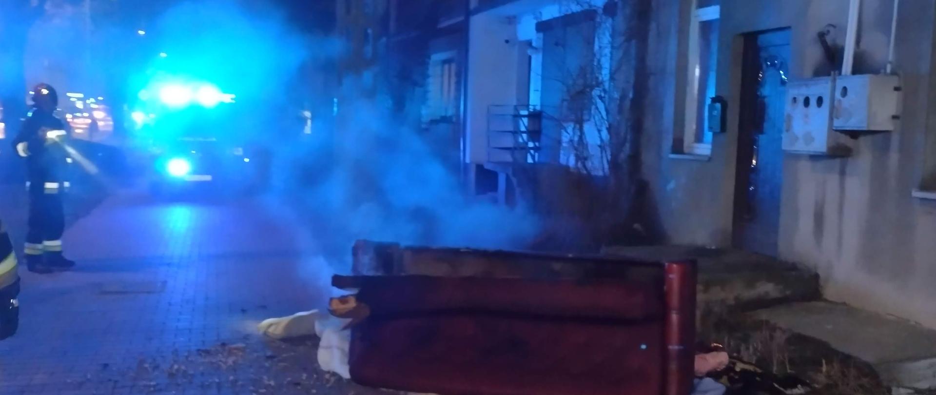 Tląca się kanapa na chodniku przed budynkiem mieszkalnym. Obok strażacy i radiowóz policyjny, panuje pora nocna. 