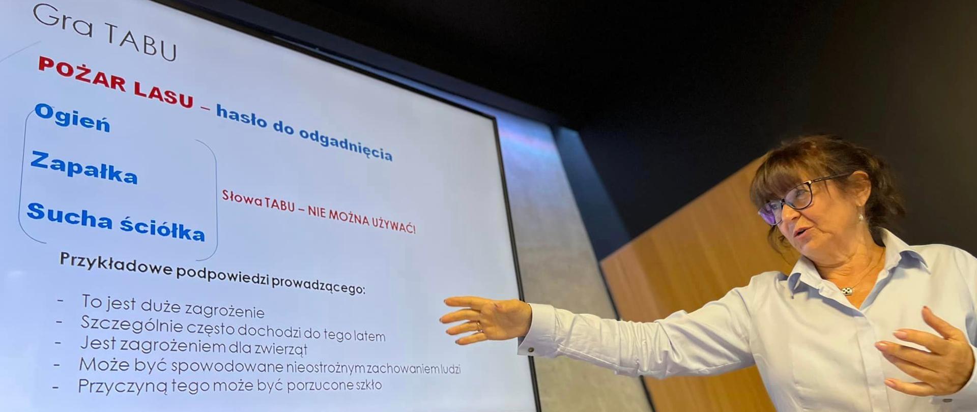 Kobieta w białej bluzce wskazującą na treść szkolenia wyświetlaną na ekranie telewizora.