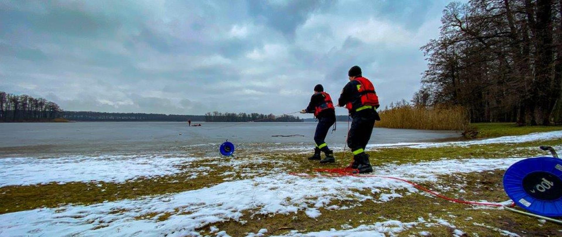 Dwóch strażaków na brzegu wyciąga linę na które podczepione są sanie lodowe. W tle strażacy na lodzie.