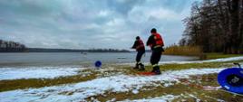 Dwóch strażaków na brzegu wyciąga linę na które podczepione są sanie lodowe. W tle strażacy na lodzie.