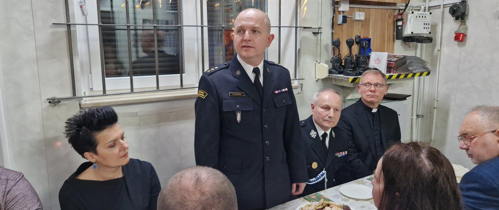 Strażak w mundurze galowym stoi i przemawia do grupy osób siedzących przy stole zastawionym słodyczami. Wśród słuchaczy znajdują się inni funkcjonariusze, ksiądz oraz cywile. Tło stanowi pomieszczenie z oknem ozdobionym światełkami i sprzętem łączności na ścianie.