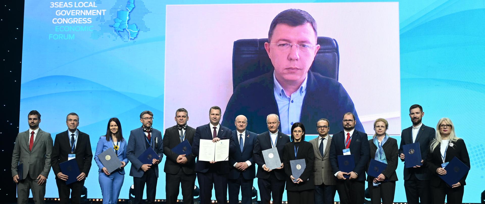 Na tle wielkiego niebieskiego ekranu na którym widać mężczyznę siedzącego na krześle stoi w szeregu grupa kilkunastu osób, mężczyzna pośrodku pokazuje otwarty dokument.