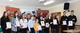 zdjęcie grupowe uczestników turnieju wiedzy pożarniczej, kilkanaście osób pozuje z pamiątkowymi dyplomami trzymając je w ręku, w tle stoją organizatorzy konkursu