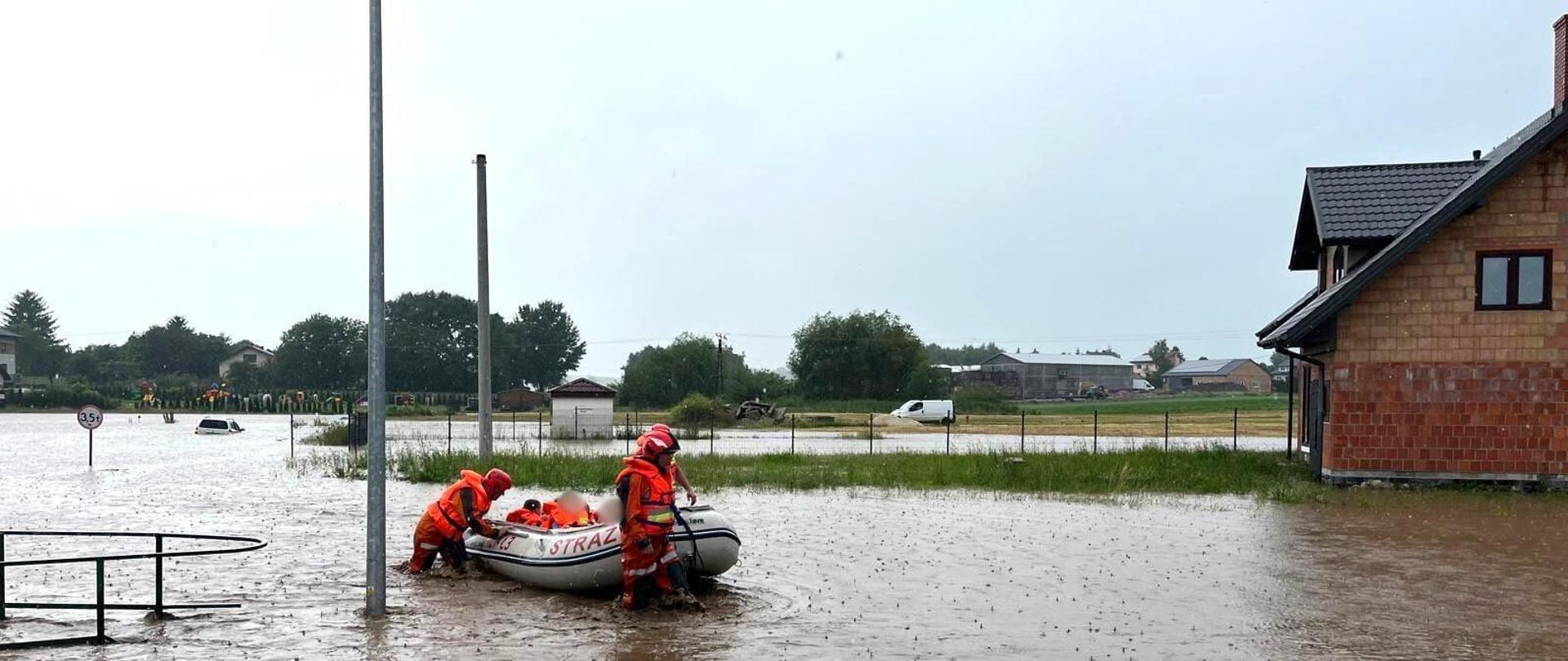 Na zdjęciu widzimy zalaną drogę po opadach i strażaków którzy przy wykorzystaniu pontonu przemieszczają się po wodzie. W tle zalany samochód po sufit.