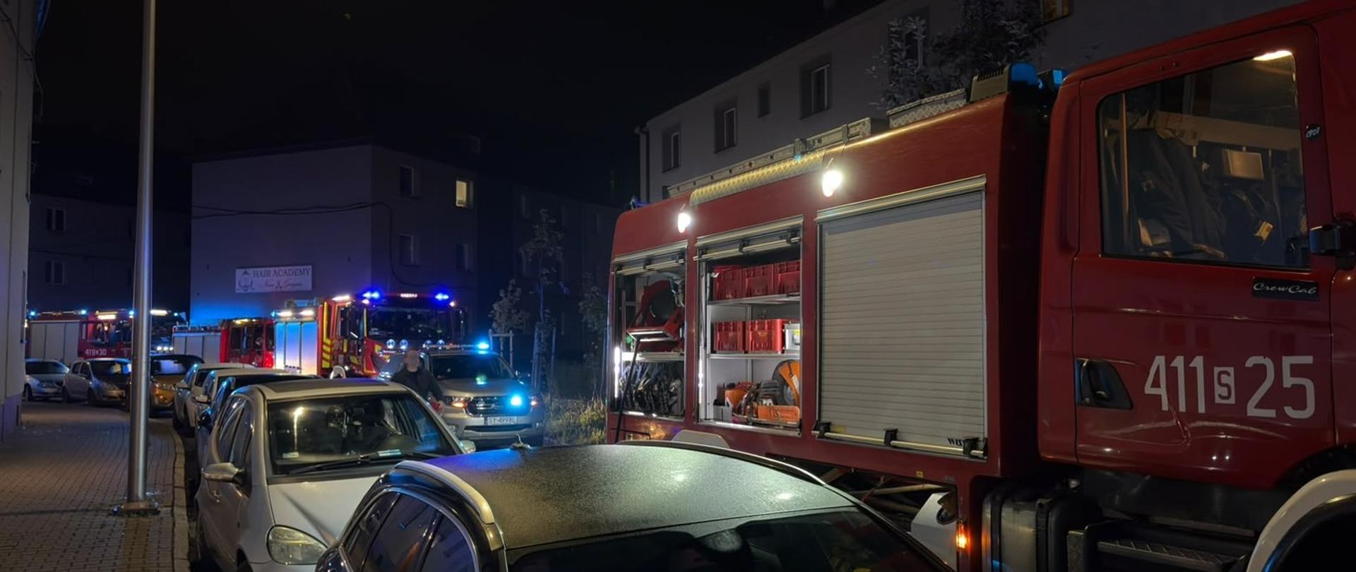 Na osiedlowej ulicy widoczny rząd wozów strażackich. Jest noc. Wszystkie auta mają włączone niebieskie światła błyskowe. Pozostałe samochody zaparkowane wzdłuż chodnika.