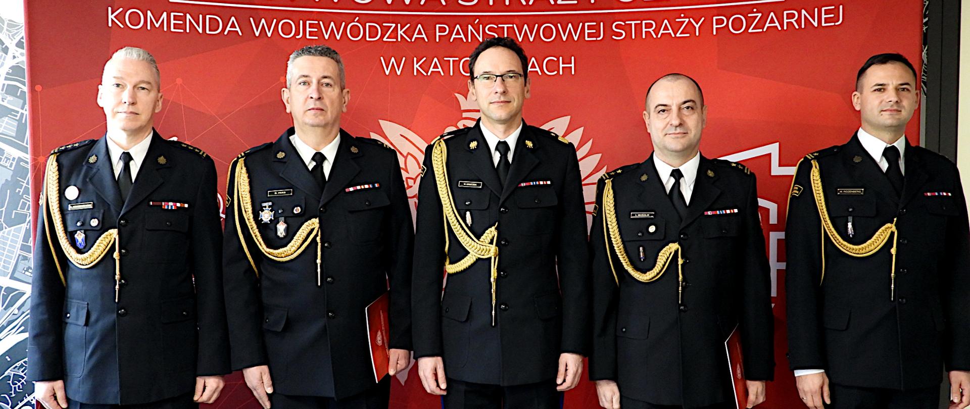 Na tle czerwonej ścianki z białym orłem w koronie po lewej stronie a po prawej logo Państwowej Straży Pożarnej. Na środku napis Państwowa Straż Pożarna. Komenda Wojewódzka PSP w Katowicach, stoją w mundurach wyjściowych, w środku śląski komendant wojewódzki nadbryg. Wojciech Kruczek, po prawej odwołany zastępca komendanta miejskiego PSP w Dąbrowie Górniczej - bryg. Łukasz Musialik i komendant miejski PSP w Dąbrowie Górniczej - mł. bryg. Mariusz Rozenberg. Po lewej stronie stoją odwołany zastępca komendanta miejskiego PSP w Katowicach mł. bryg. Dariusz Paks i komendant miejski PSP w Katowicach st. bryg. Arkadiusz Korzeniewski. 