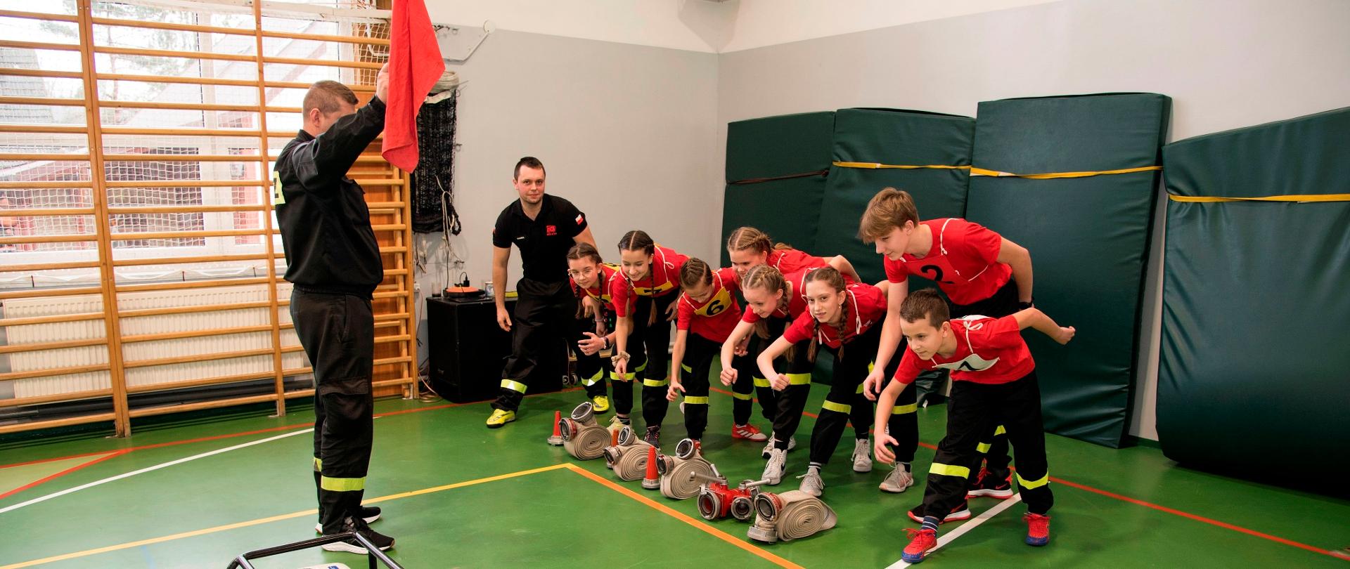 Widok z boku. Drużyna w pozycji startowej do rozpoczęcia konkurencji. Po lewej stronie sędzia startowy z chorągiewką.