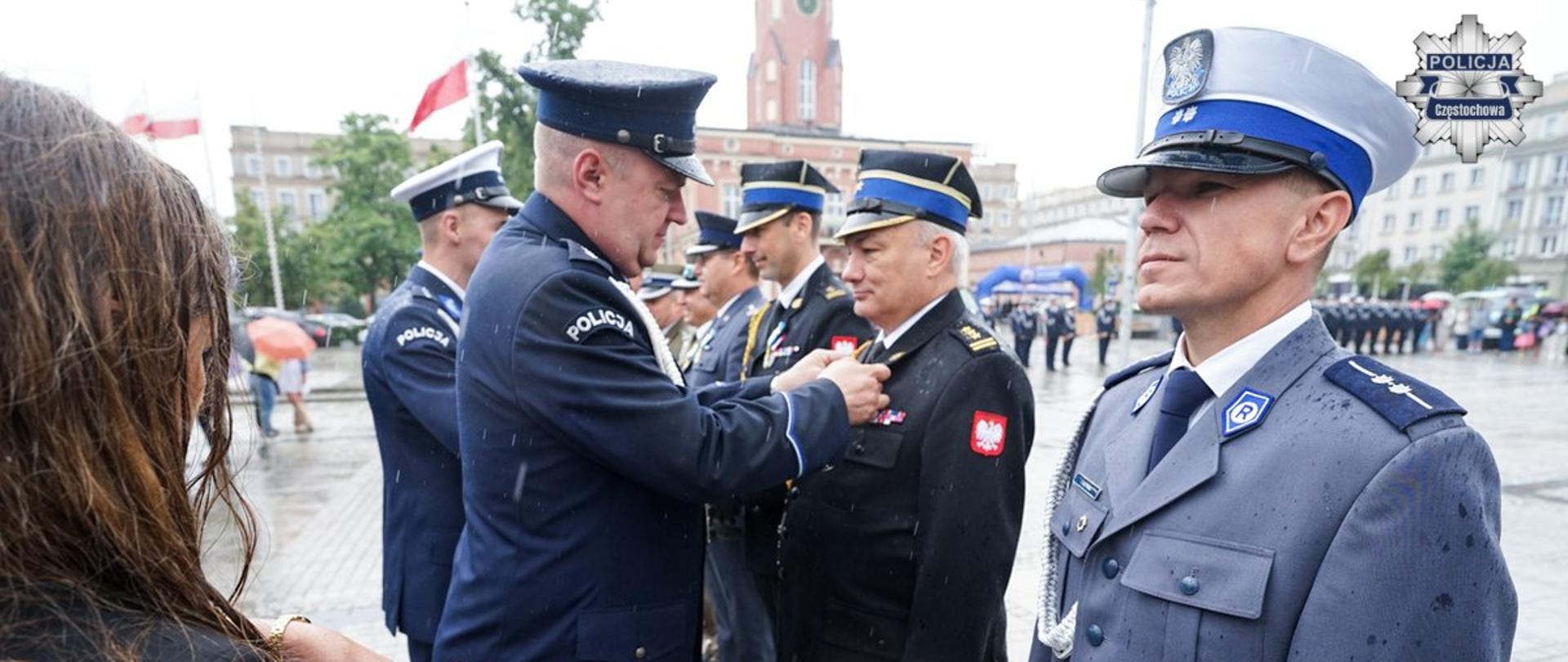 Funkcjonariusz Policji przypina do klapy munduru medal Komendantowi Centralnej Szkoły PSP, po lewej kobieta trzymająca tacę pokrytą czerwonym suknem, na której leżą medale