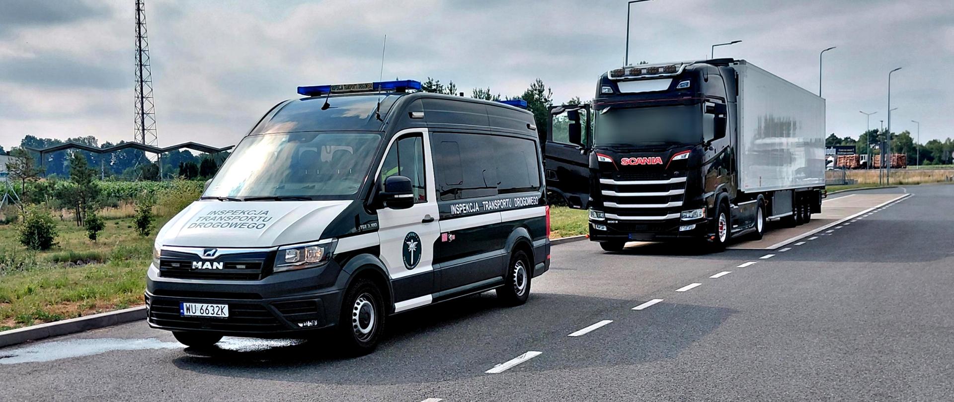 Samochód Inspekcji Transportu Drogowego z włączonymi niebieskimi światłami stoi przed czarnym ciągnikiem siodłowym Scania z białą naczepą chłodniczą na poboczu szerokiej drogi. W tle widoczne są pola, niska zabudowa oraz pochmurne niebo.