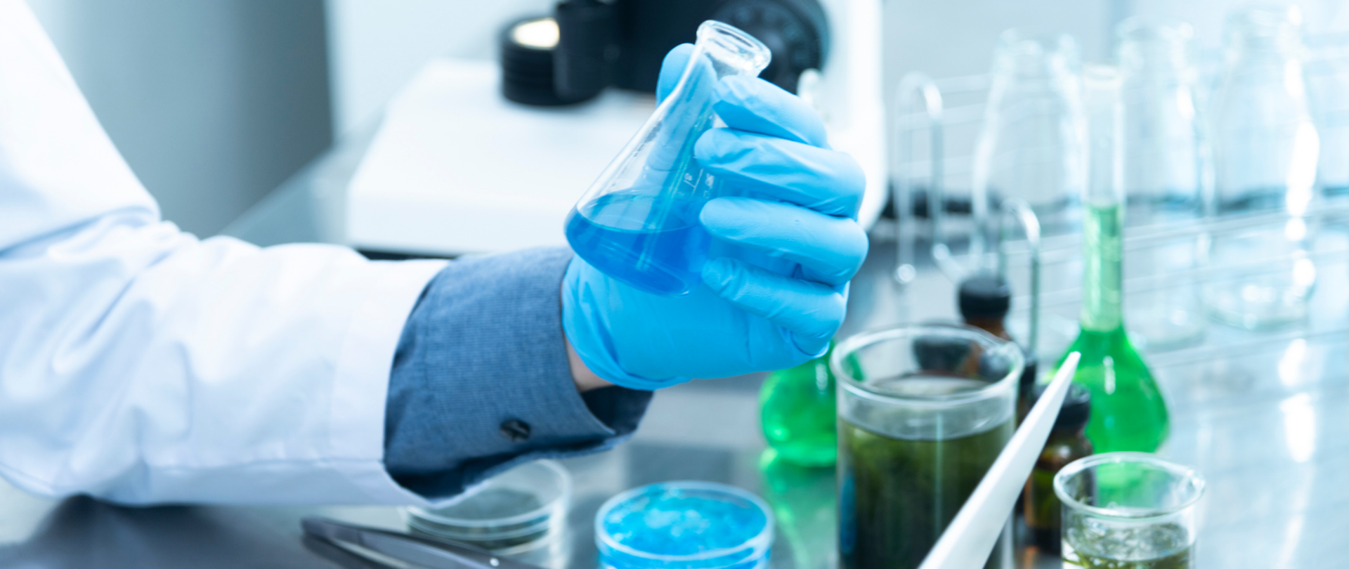 Laboratorium - ręka w białym kitlu i niebieskiej, gumowanej rękawiczce trzyma fiolkę wypełnioną niebieskim płynem. Obok próbki z zielonymi i niebieskimi płynami, a także puste fiolki. W tle mikroskop.