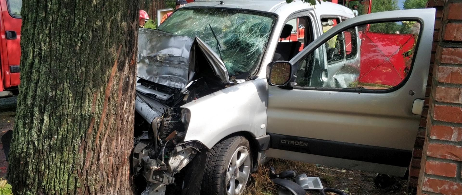 Na zdjęciu widoczny samochód osobowy Citroen Berlingo koloru srebrnego rozbity o przydrożne drzewo. Na drugim planie samochody straży pożarnej.