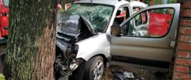 Na zdjęciu widoczny samochód osobowy Citroen Berlingo koloru srebrnego rozbity o przydrożne drzewo. Na drugim planie samochody straży pożarnej.