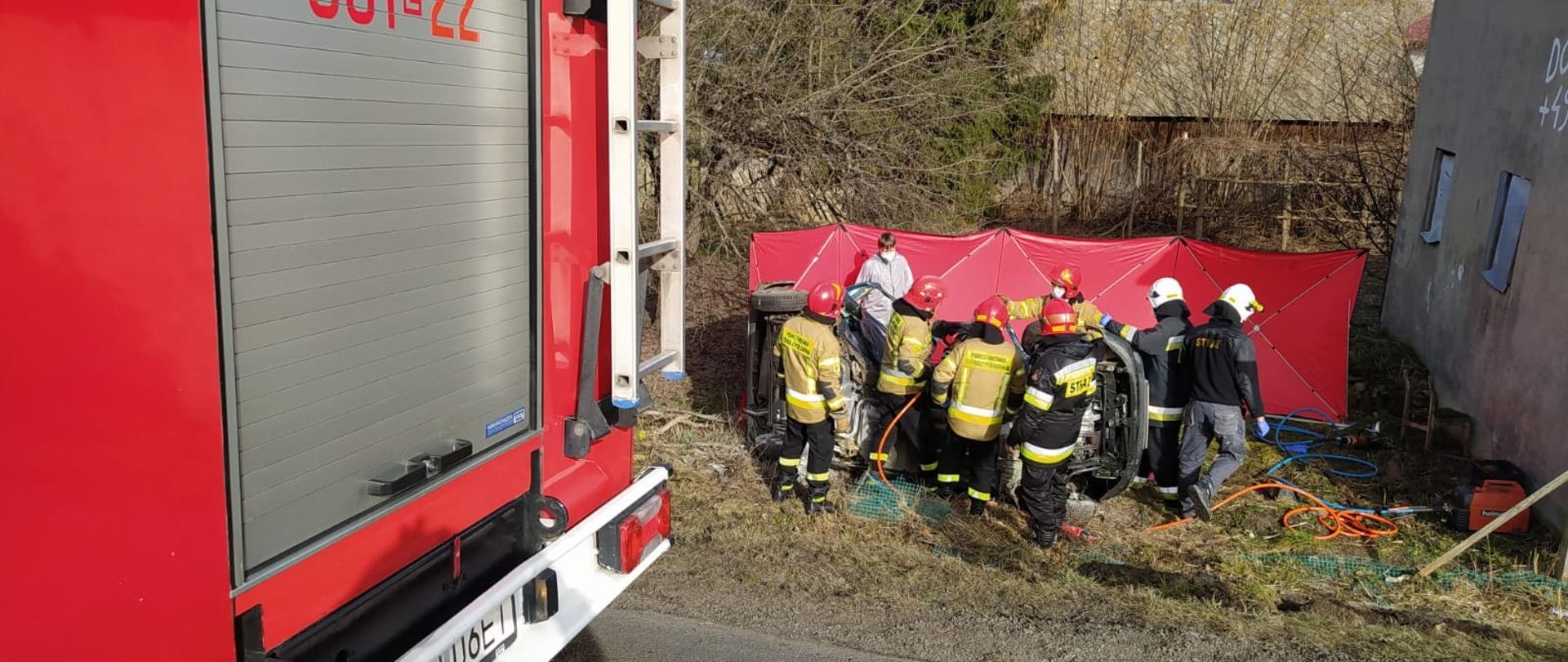 W odległości ok 3 metrów od jezdni w dole lezy na boku samochód , za min rozstawiony jest czerowny parawan. Działania przy nim wykonuje siedmiu strażaków