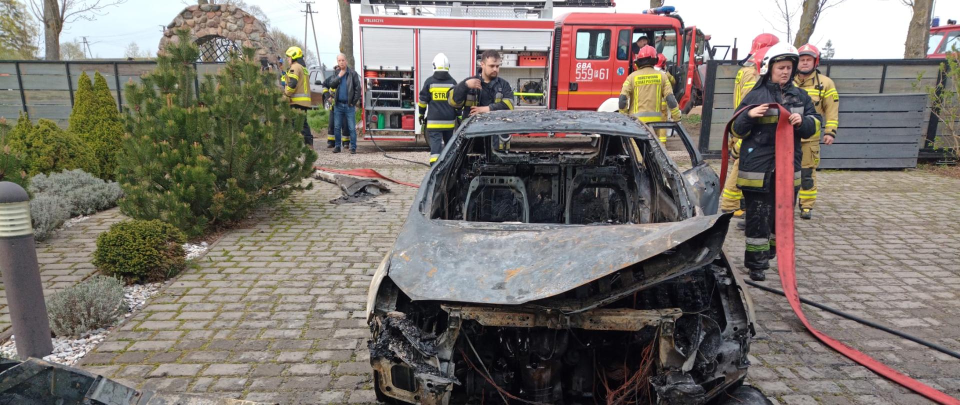 Na zdjęciu widać spalony samochód osobowy w tle strażacy zwijający sprzęt oraz samochód pożarniczy GBA..