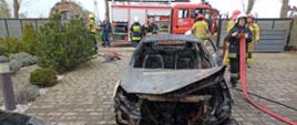 Na zdjęciu widać spalony samochód osobowy w tle strażacy zwijający sprzęt oraz samochód pożarniczy GBA..