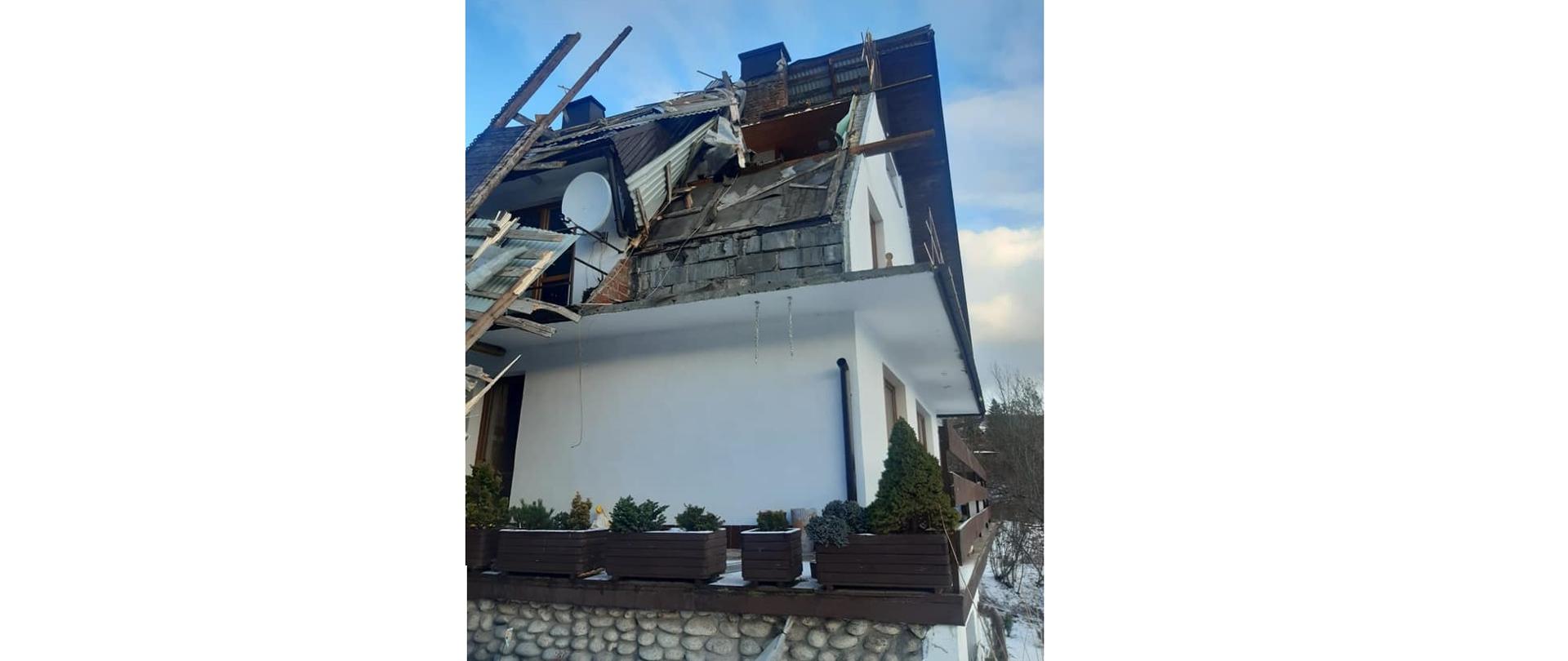 Zdjęcie przedstawia zerwany dach w Kościelisku po przejściu wiatru halnego. Na pierwszym planie widać zerwany dach budynku, w tle drzewa i pochmurne niebo.