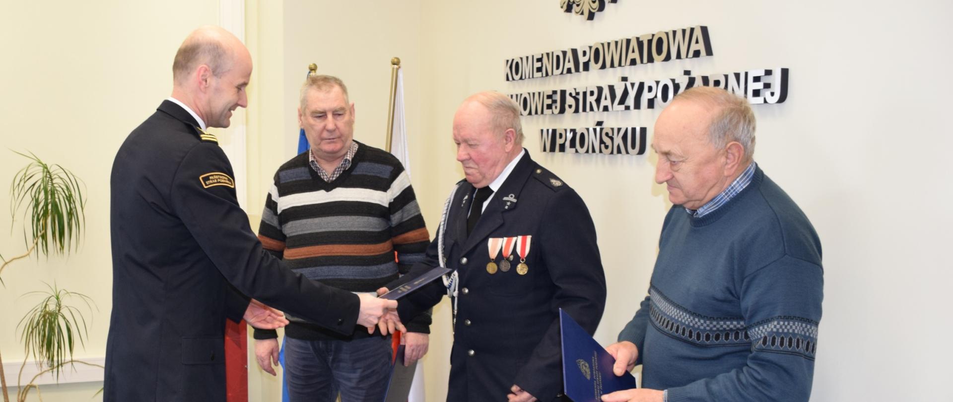 Oficer Państwowej Straży Pożarnej, w granatowym mundurze wręcza dokument strażakowi OSP w mundurze wyjściowym, w kolorze granatowym, z trzema odznaczeniami. Obok wręczanego, z prawej i lewej strony stoją dwie osoby w ubraniach cywilnych - swetrach i ciemnych spodniach. 