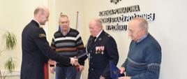 Oficer Państwowej Straży Pożarnej, w granatowym mundurze wręcza dokument strażakowi OSP w mundurze wyjściowym, w kolorze granatowym, z trzema odznaczeniami. Obok wręczanego, z prawej i lewej strony stoją dwie osoby w ubraniach cywilnych - swetrach i ciemnych spodniach.