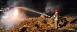 Na zdjęciu widać strażaka w czerwonych hełmie i żółtym ubraniu, który leje wodę na palącą się słomę. W akcji pomaga mu traktor z przyczepionymi do podnośnika widłami. W tle widać zarośla i drzewo. Jest noc.