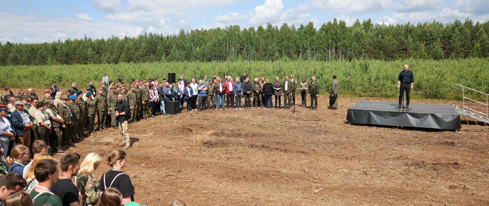 Na tle lasu ludzie stoją w półokręgu. Po prawej stronie na podwyższeniu stoi mężczyzna ubrany w ciemną koszule i spodnie. 