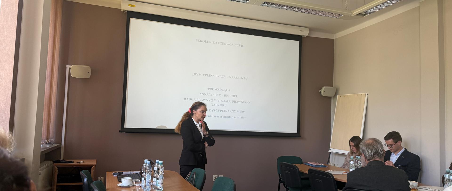 Prowadząca szkolenie prezentuje uczestnikom zagadnienia związane z tematyką wystąpienia. w tle widoczna jest prezentacja multimedialna wyświetlana na ekranie.