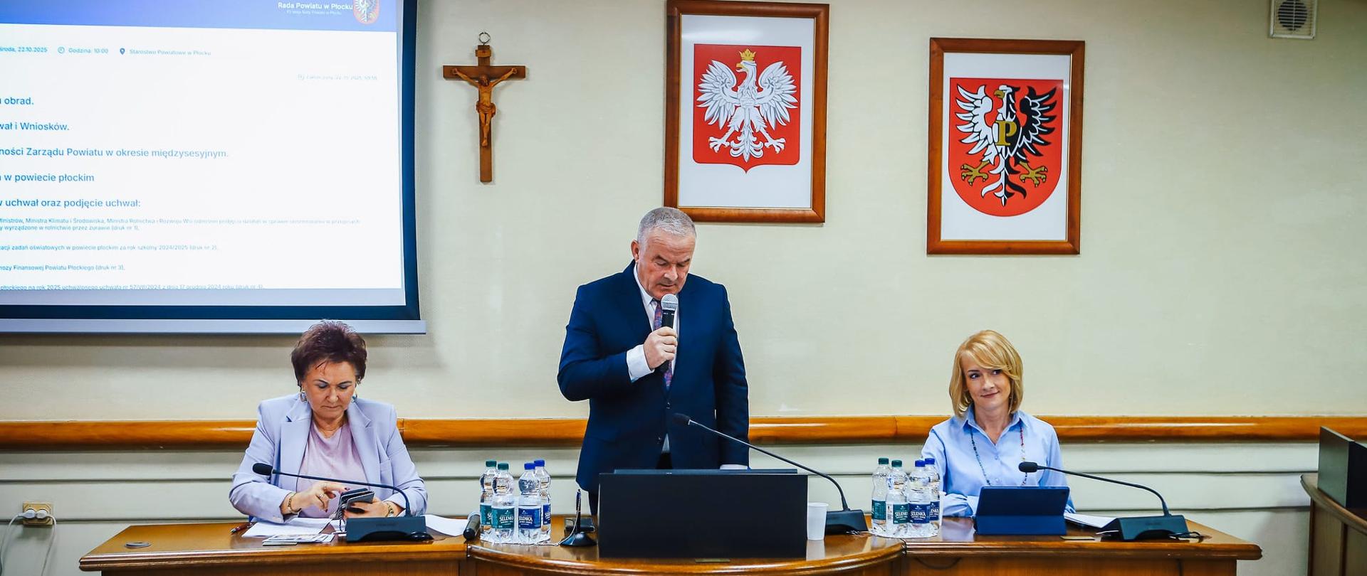 Formalne posiedzenie w sali obrad. Trzy osoby siedzą przy stole prezydialnym, a mężczyzna na środku przemawia do mikrofonu. Na ścianie za nimi wiszą godła oraz ekran z programem obrad. Atmosfera urzędowa i oficjalna.