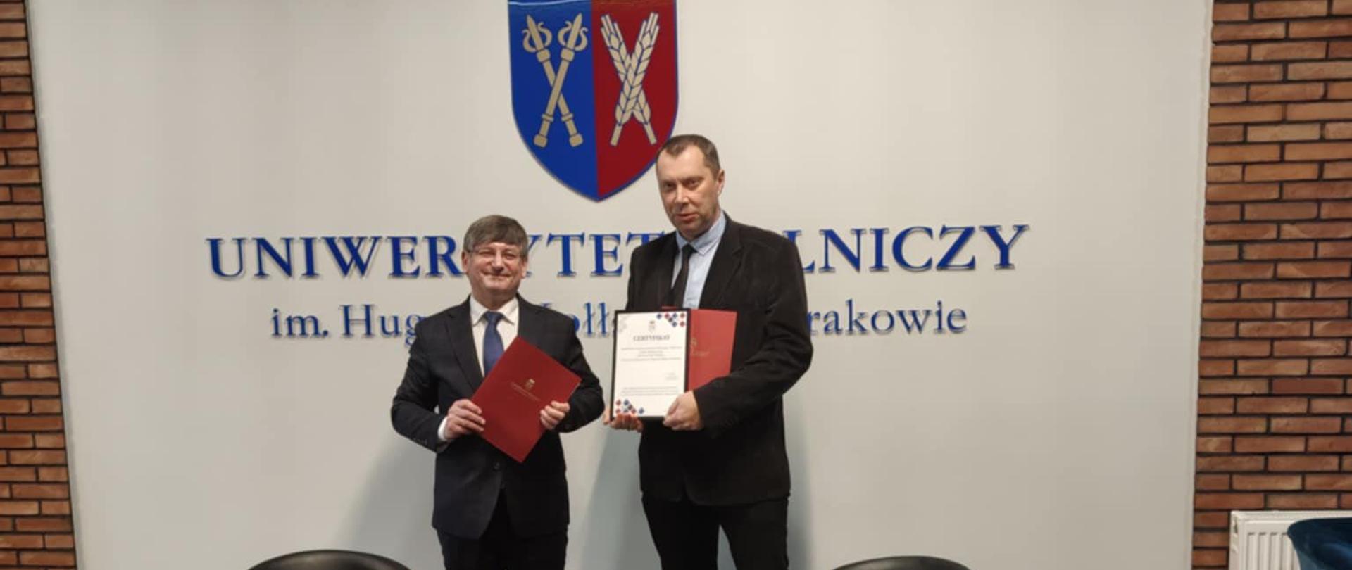 Dwaj mężczyźni stoją przed ścianą z herbem i napisem „Uniwersytet Rolniczy w Krakowie”; jeden trzyma teczkę, drugi prezentuje certyfikat szkoły partnerskiej.