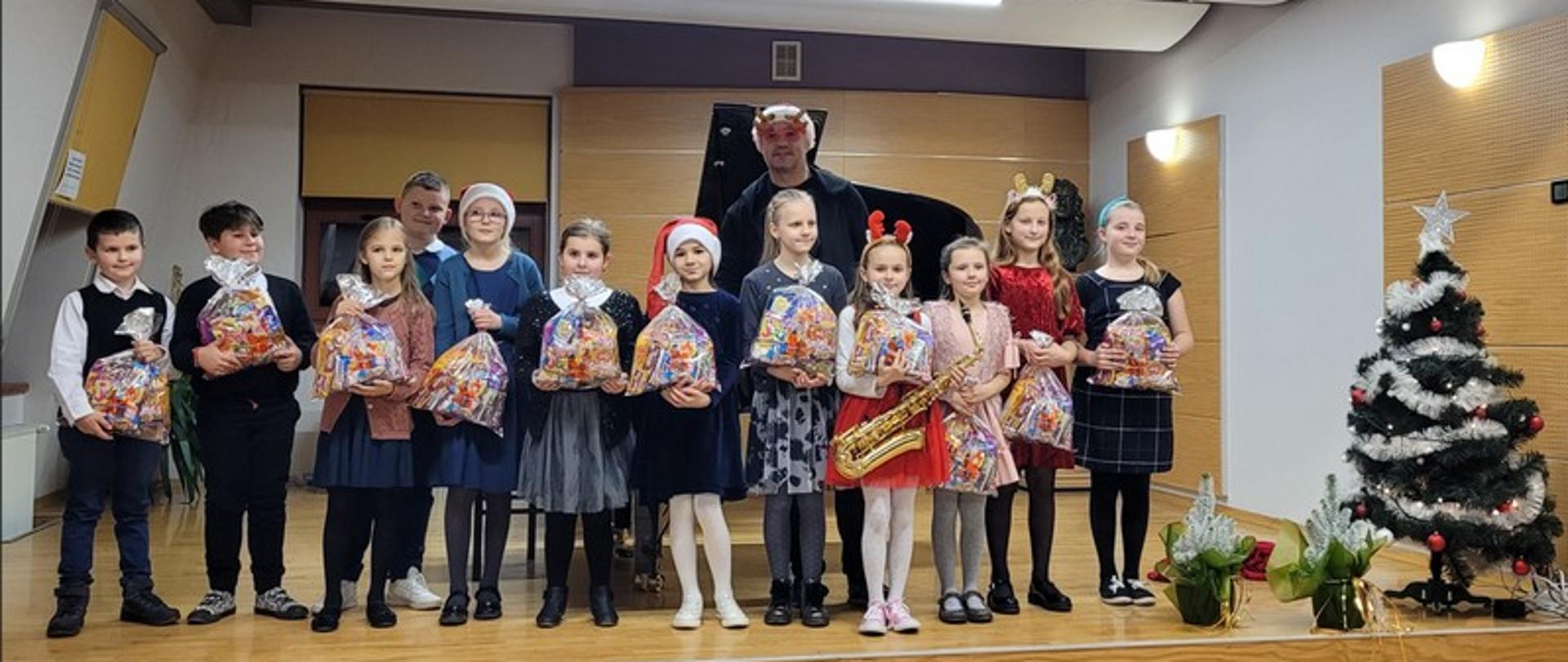 Zdjęcie przedstawia uczestników audycji Mikołajkowej stojących na scenie wraz z otrzymanymi paczkami mikołajkowymi. Za dziećmi stoi nauczyciel w czapce Mikołaja oraz okularach w renifery. Po prawej stroni zdjęcia choinka oraz dwie małe choinki.