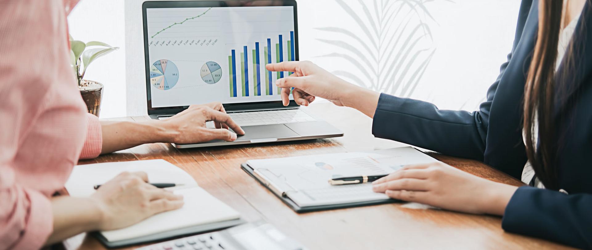 Dwie osoby przy biurku, jedna pokazuje na ekran komputera.