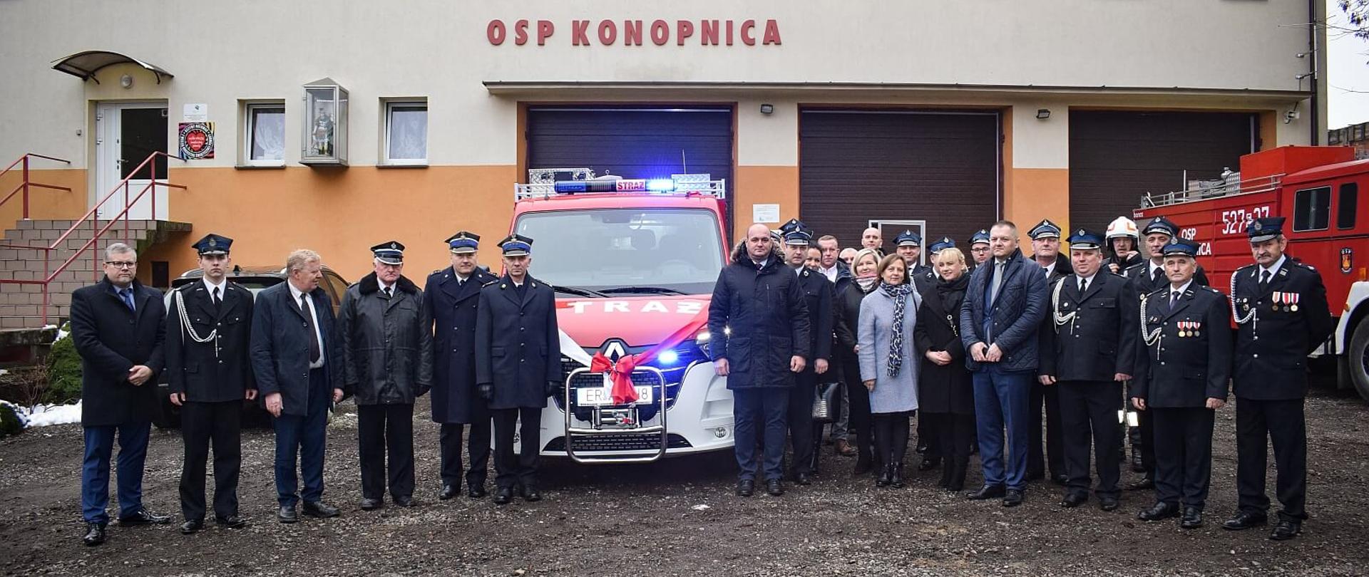 uczestnicy uroczystości przekazania samochodu pozują do zdjęcia przed budynkiem osp, wśród zebranych są strażacy psp druhowie osp, politycy oraz przedstawiciele gminy i związku osp 