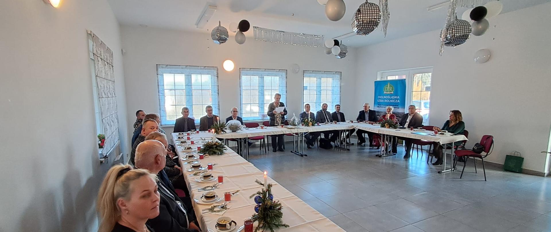 Zdjęcie przedstawia posiedzenie rady w jasnej, przestronnej sali. Kilkanaście osób siedzi przy długich stołach ustawionych w kształcie podkowy, nakrytych białymi obrusami. Na stołach znajdują się filiżanki, talerze z poczęstunkiem, szklanki z napojami oraz świąteczne dekoracje z gałązek i świec. W centralnej części sali jedna osoba stoi i przemawia do uczestników. W tle widoczny jest baner Dolnośląskiej Izby Rolniczej oraz duże okna wpuszczające światło dzienne. Sufit ozdobiony jest balonami i dekoracjami, a uczestnicy spotkania są ubrani w stroje formalne.