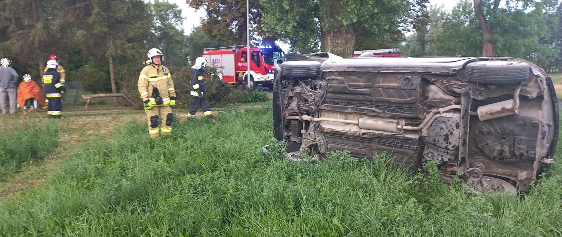 Po prawej stronie leży samochód osobowy na prawym boku. Widać podwozie auta. Po środku na drodze stoi samochód pożarniczy Z lewej strony siedzi osoba poszkodowana. Obok niej członek rodziny i strażacy