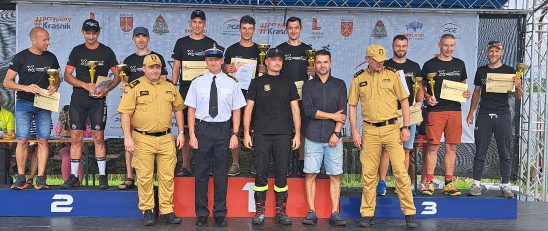 Grupa sportowców oraz strażacy w mundurach służbowych stoją na scenie i podium