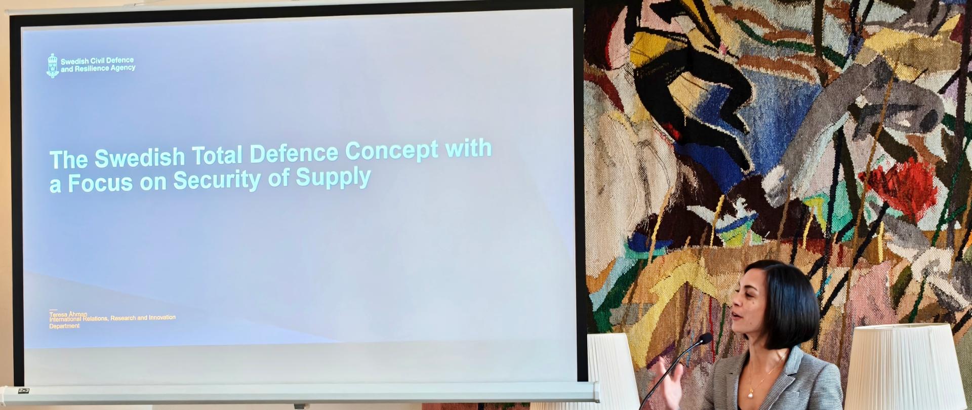 Prelegentka stoi przy mównicy i wygłasza prezentację w sali konferencyjnej. Obok niej znajduje się ekran z prezentacją zatytułowaną „The Swedish Total Defence Concept with a Focus on Security of Supply”. Uczestnicy siedzą przy stołach i słuchają wystąpienia.