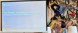 Prelegentka stoi przy mównicy i wygłasza prezentację w sali konferencyjnej. Obok niej znajduje się ekran z prezentacją zatytułowaną „The Swedish Total Defence Concept with a Focus on Security of Supply”. Uczestnicy siedzą przy stołach i słuchają wystąpienia.