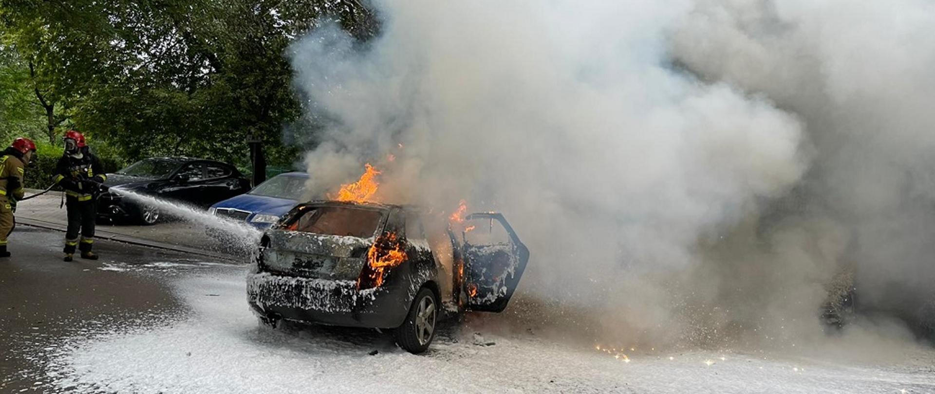 Na zdjęciu po jego lewej stronie widać dwóch funkcjonariuszy Straży Pożarnej podających prąd piany ciężkiej. W centralnej części zdjęcia widać samochód koloru czarnego z którego wnętrza wydobywają się płonienie oraz gęste zadymienie.