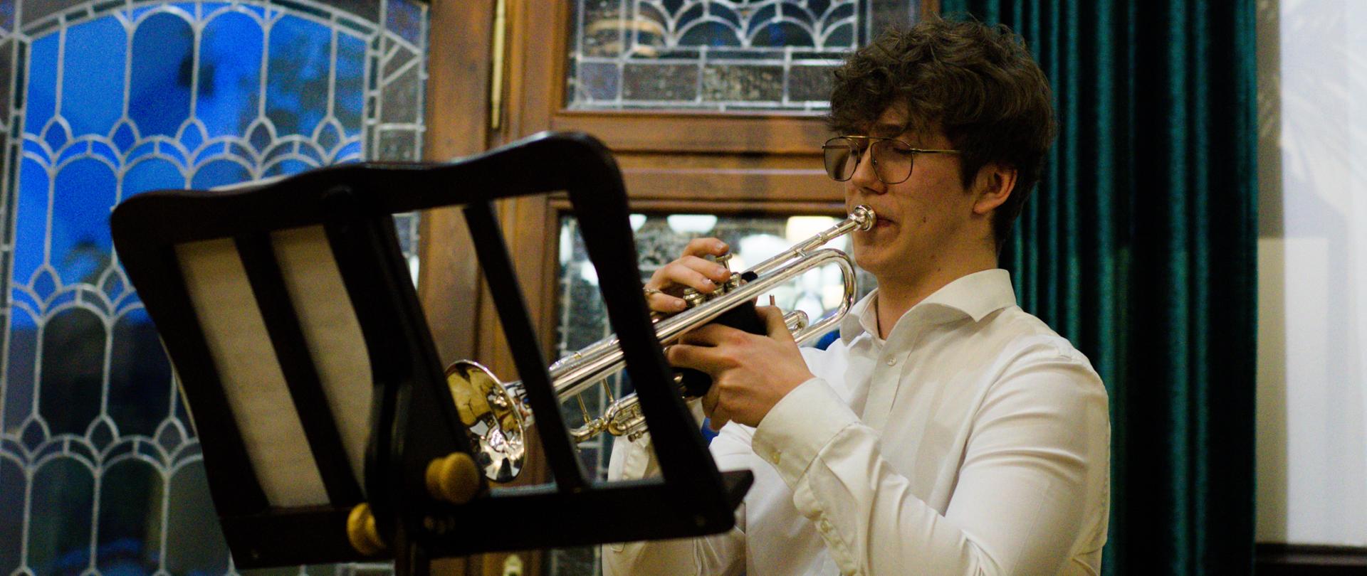 Mody mężczyzna w białej koszuli i okularach gra na trąbce. Przed nim stoi pulpit, na którym znajdują się nuty. W tle okno z witrażem i zielone zasłony.