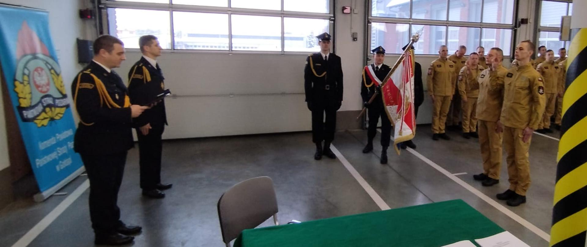 Zdjęcie przedstawia uroczysty moment składania ślubowania przez nowo przyjętych funkcjonariuszy strażaka Krzysztofa Pojawy oraz strażaka Adama Sokołowskiego do służby w Komendzie Powiatowej Państwowej Straży Pożarnej w Gołdapi oraz sztandar komendy. Na obrazie można również zaobserwować pozostałych strażaków biorących udział w uroczystej zbiórce