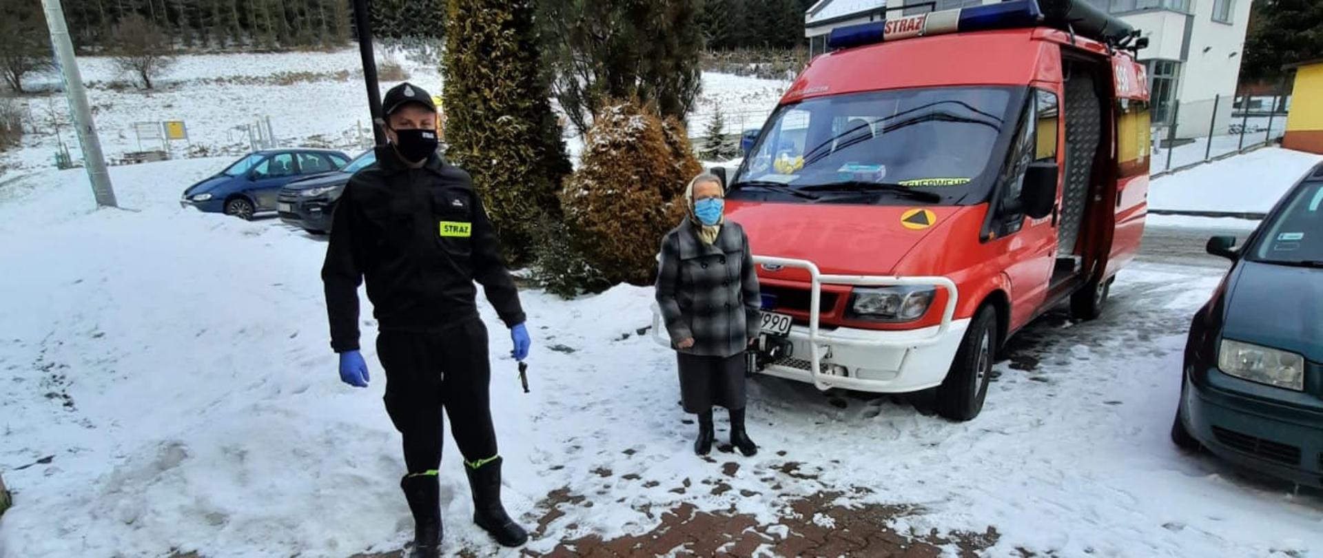 Zdjęcie przedstawia strażaka i seniora na tle samochodu strażackiego.