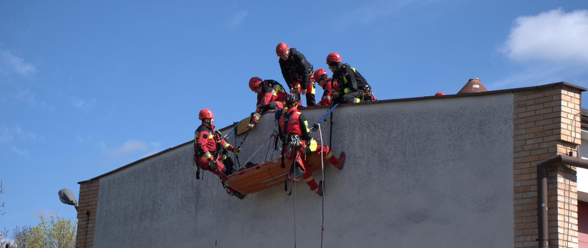 Ratownicy na dachu budynku przygotowują ewakuację poszkodowanego w noszach opuszczanych na linach.
