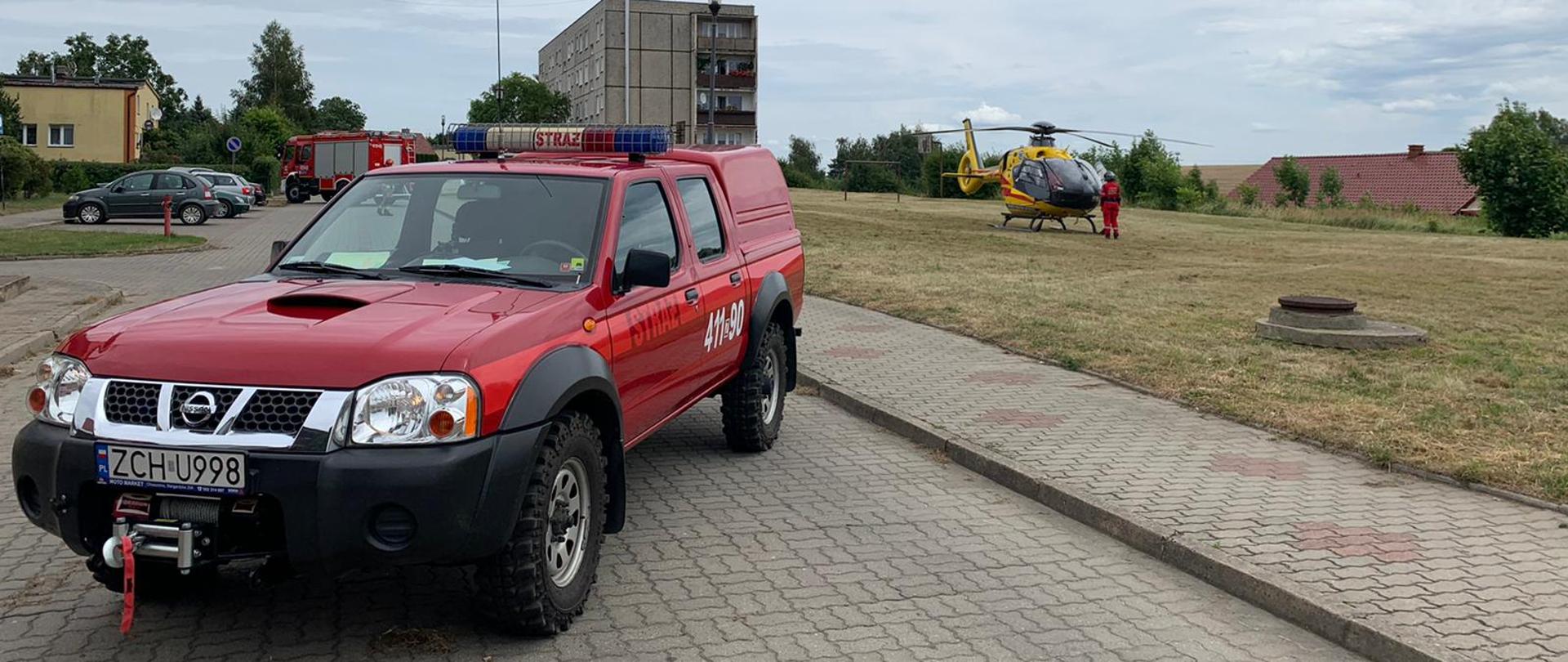 Na fotografii po lewej stronie, na drodze znajdują się zaparkowane w poprzek dwa wozy strażackie. Pomiędzy wozami, na parkingu po lewej stronie stoją zaparkowane pojazdy. Po prawej stronie na boisku wylądował śmigłowiec Lotniczego Pogotowia Ratunkowego. Przed śmigłowcem stoi ratownik medyczny. W oddali znajduje się blok oraz inne budynki i drzewa. Zdjęcie wykonano w porze dziennej.
