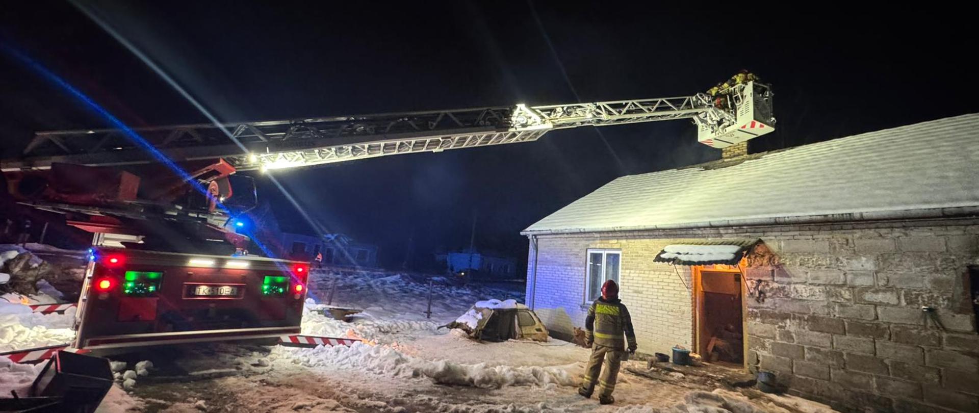 Strażacy na podnośniku w porze nocnej zaglądają do komina domu mieszkalnego.