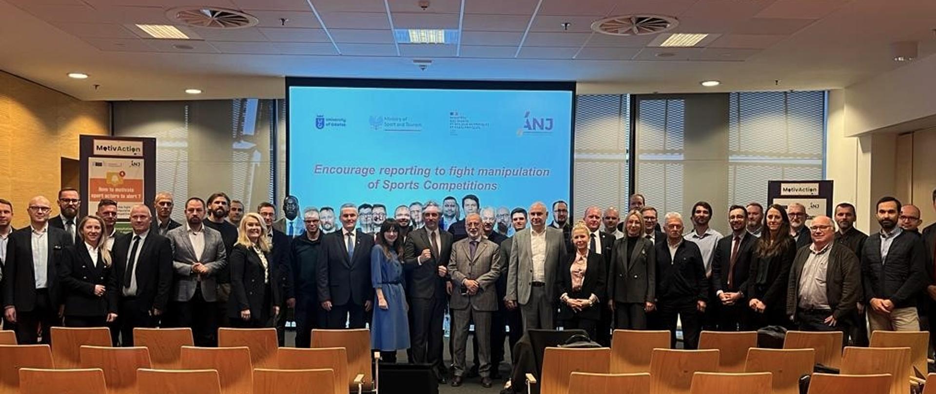 Zdjęcie przedstawia liczną grupę osób, która stoi na tle slajdu z angielskim tytułem konferencji naukowej, którym jest: wspierania zgłaszania nieprawidłowości w celu zwalczania manipulowania zawodami sportowymi.