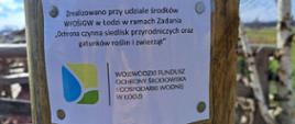 do słupa przyczepiona jest biała tablica z napisem zrealizowano przy udziale środków WFOŚiGW w Łodzi w ramach Zadania ,,Ochrona czynna siedlisk przyrodniczych oraz gatunków roślin i zwierząt" poniżej logo oraz napis Wojewódzki Fundusz Ochrony Środowiska i Gospodarki Wodnej w Łodzi 