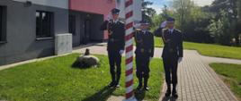 Trzech strażaków stoi za masztem flagowym, wszyscy salutują. Za nimi w tle ściana budynku Komendy w kolorach szaro - czerwonych i napisem Komenda Miejska Państwowej Straży Pożarnej w Żorach. W prawej części zdjęcia w tle drzewa. 