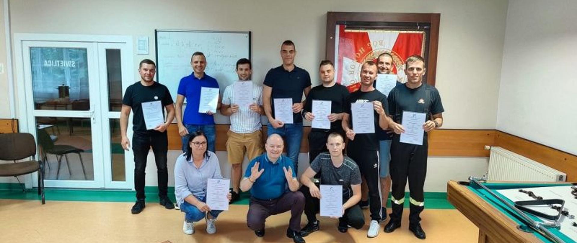 Zdjęcie wykonane na świetlicy KP PSP Białogard. Zdjęcia przedstawia osoby, które ukończyły kurs wraz z osobą prowadzącą. W tle sztandar KP.Osoby trzymają dyplomy potwierdzające zakończenie kursu.